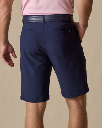 Men's Maverick Twill Short in New Indigo, back model view featuring a navy solid twill fabric with buttoned welt back pockets and tonal logo detail, styled with a pink polo and navy belt