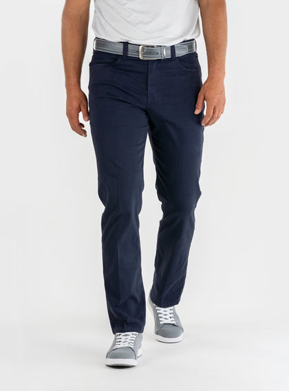 Men's Clemson Pant in Indigo, on-model front view featuring a navy five-pocket style with a button closure and gray belt worn with a white tee and gray golf shoes