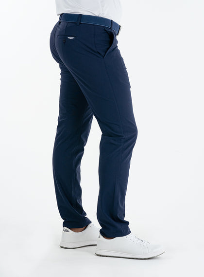 Men's Quest Pant in Indigo, side and back on-model view featuring navy pants with a small embroidered logo on the back pocket, styled with a white shirt and white shoes