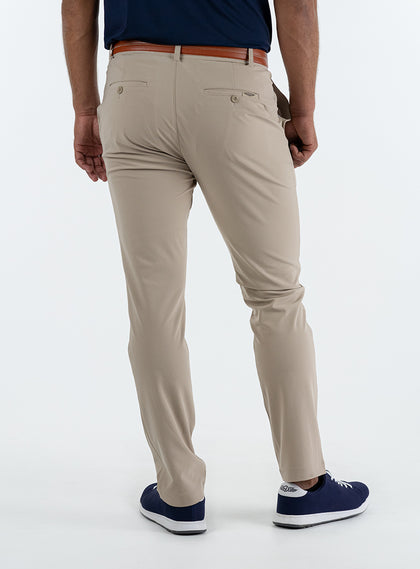 Men's Quest Pant in Light Khaki, back on-model view featuring khaki pants with button-closure back pockets and a small embroidered logo on back pocket, styled with a navy shirt, brown belt, and navy shoes