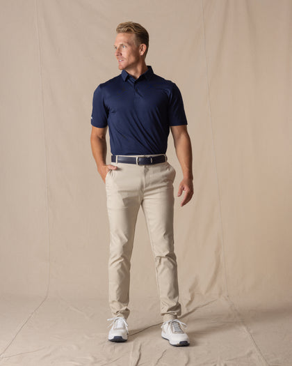 Men's Maverick Twill Pant in Cashew, full length front view on model featuring a solid cashew color paired with a navy polo, navy belt, and white golf shoes