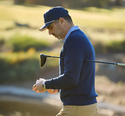 A man on a golf course wearing a navy blue long-sleeve sweater, khaki pants, and a navy cap, holding a golf club over his shoulder while looking down at his watch.