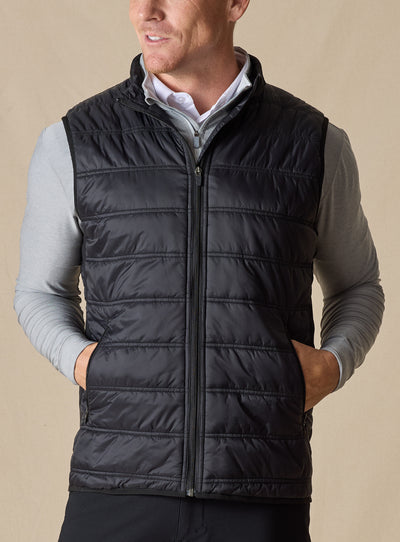 Men's Delta Vest in Black, front model view featuring horizontal quilted baffles and full-zip closure, layered over a gray long-sleeve pullover and white button-down shirt, styled with black pants
