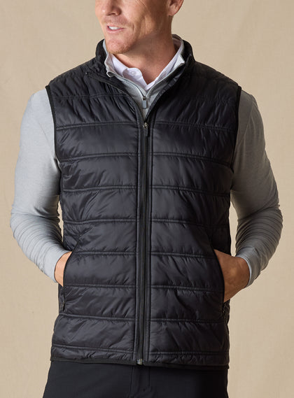 Men's Delta Vest in Black, front model view featuring horizontal quilted baffles and full-zip closure, layered over a gray long-sleeve pullover and white button-down shirt, styled with black pants
