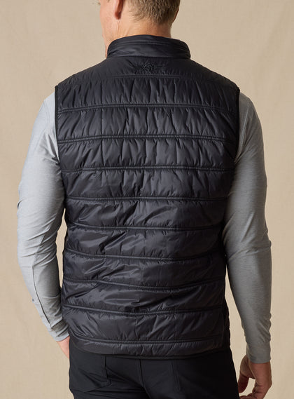 Men's Delta Vest in Black, back model view featuring horizontal quilted baffles and tonal embroidered winged logo at the upper back yoke, layered over a gray long-sleeve pullover, styled with black pants