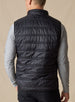 Men's Delta Vest in Black, back model view featuring horizontal quilted baffles and tonal embroidered winged logo at the upper back yoke, layered over a gray long-sleeve pullover, styled with black pants