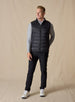 Men's Delta Vest in Black, full-length front model view featuring horizontal quilted baffles and full-zip closure, layered over a gray long-sleeve pullover, styled with black pants and white golf shoes against a neutral canvas backdrop