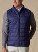 Men's Delta Vest in Indigo, front model view featuring horizontal quilted baffles and full-zip closure, layered over a gray long-sleeve pullover and white button-down shirt, styled with white pants