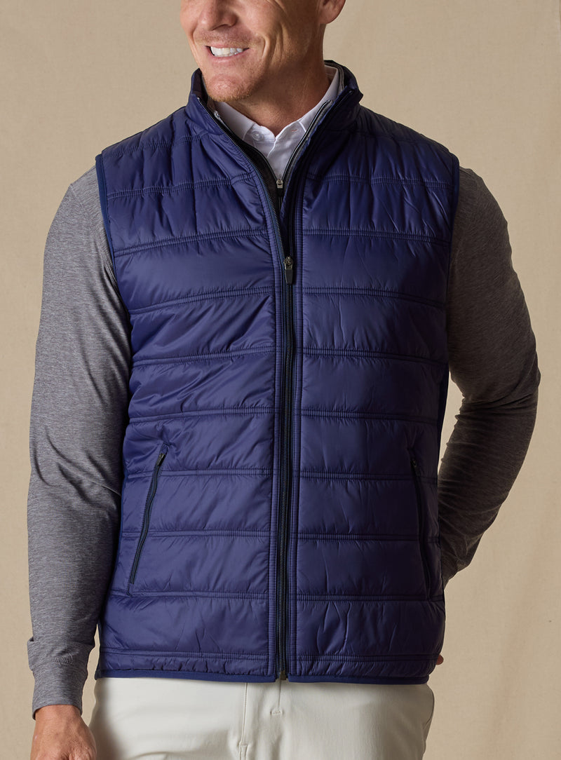 Men's Delta Vest in Indigo, front model view featuring horizontal quilted baffles and full-zip closure, layered over a gray long-sleeve pullover and white button-down shirt, styled with white pants