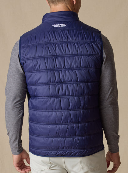 Men's Delta Vest in Indigo, back model view featuring horizontal quilted baffles and embroidered winged logo at the upper back yoke, layered over a gray long-sleeve pullover, styled with white pants