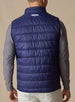 Men's Delta Vest in Indigo, back model view featuring horizontal quilted baffles and embroidered winged logo at the upper back yoke, layered over a gray long-sleeve pullover, styled with white pants