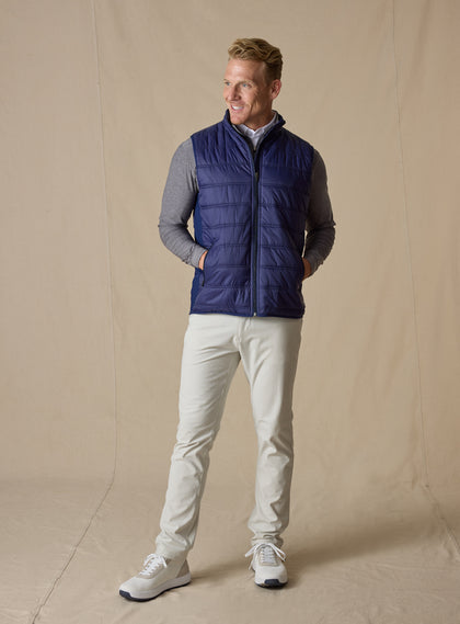 Men's Delta Vest in Indigo, full-length front model view featuring horizontal quilted baffles and full-zip closure, layered over a gray long-sleeve pullover, styled with stone pants and white shoes against a neutral canvas backdrop