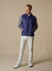 Men's Delta Vest in Indigo, full-length front model view featuring horizontal quilted baffles and full-zip closure, layered over a gray long-sleeve pullover, styled with stone pants and white shoes against a neutral canvas backdrop