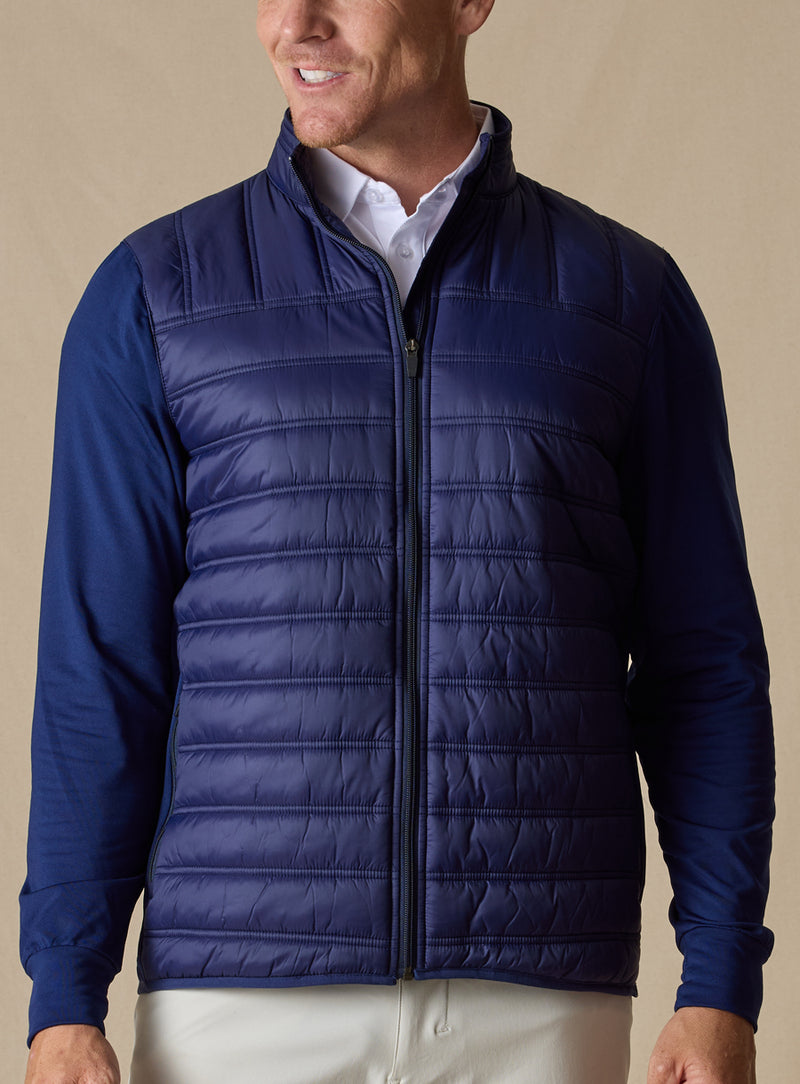 Men's Patton Jacket in Indigo, front on-model view featuring a navy quilted hybrid jacket with a zip front, quilted puffer body, and smooth stretch knit sleeves, styled with white pants
