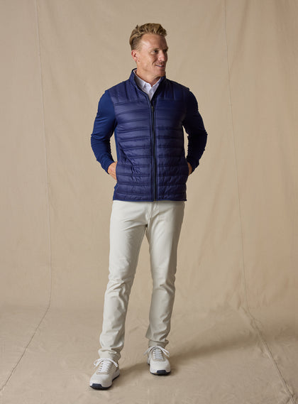 Men's Patton Jacket in Indigo, full-length on-model view featuring a navy quilted hybrid jacket with a zip front, quilted puffer body, and smooth stretch knit sleeves, styled with white pants and white shoes