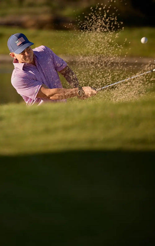 A golfer in a del mar punch polo swings a sand wedge in a bunker, kicking up sand near a water hazard.