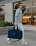 Straight Down Montana Duffle Bag in New Indigo, lifestyle model view of a man carrying the navy duffle bag by the handles while walking outdoors in a gray quarter-zip and white pants