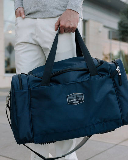 Straight Down Montana Duffle Bag in New Indigo, close-up model view highlighting the Straight Down mountain badge logo patch and dual carry handles of the navy nylon duffle bag