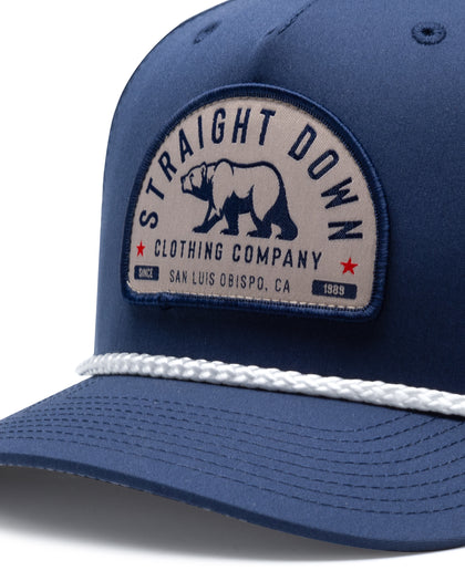 Bear Country Rope Hat in New Indigo, close-up front detail view featuring a woven bear patch reading "Straight Down Clothing Company, San Luis Obispo, CA Since 1989" with red star accents on the navy front panel with a white rope brim detail