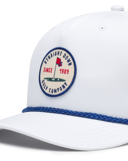 Pin High Rope Hat in White, close-up detail of the Straight Down Golf Company circular patch depicting a golf hole with a red flag and "Since 1989" text, with a blue rope brim detail