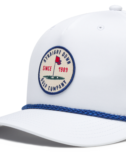 Pin High Rope Hat in White, close-up detail of the Straight Down Golf Company circular patch depicting a golf hole with a red flag and "Since 1989" text, with a blue rope brim detail