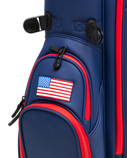 Straight Down Lux Carry Stand Bag in Navy Red, close-up detail view of lower front pocket section featuring embroidered American flag patch, red zipper trim, black zipper pulls, and navy blue structured pockets