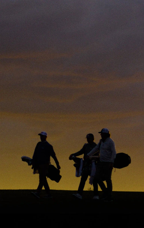 Three golfers walking across a golf course at dusk, silhouetted against a glowing sunset sky.