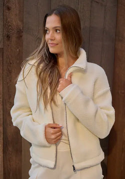 A woman wearing a cream-colored sherpa fleece zip-up jacket and matching sweatpants, standing against a rustic dark wood plank wall.