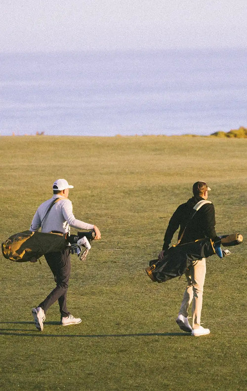 Two golfers walking across a grassy golf course at sunset, each carrying a black golf bag with clubs.