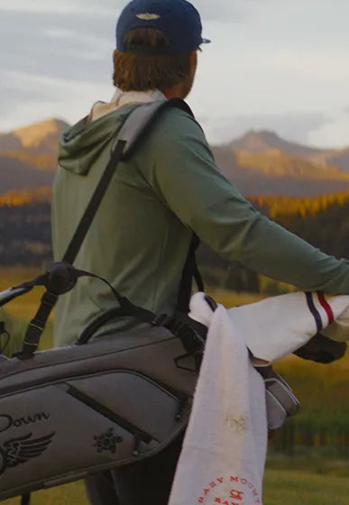 A person seen from behind wearing a green hooded long-sleeve top and a navy cap, carrying a gray golf bag with a white towel over their shoulder in an outdoor mountain landscape at sunset.
