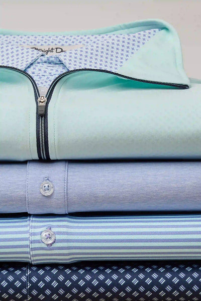 A stack of folded dress shirts in various blue and light blue patterns, including solid, stripes, and small dots, with a mint green shirt with a zipper and a patterned inner collar.