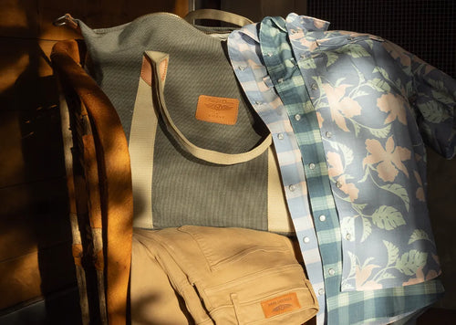 A styled flat-lay of multiple apparel items from the same brand, including a gray ribbed tote bag, a light blue floral button-up shirt, a blue-and-white plaid shirt, tan shorts, and a brown leather belt, arranged together on a dark surface with dramatic warm sunlight and shadows.