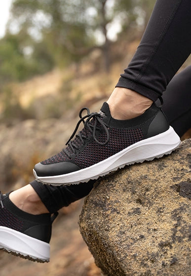 Close-up of woman wearing black Swing shoe with white soles, resting foot on a rock outdoors