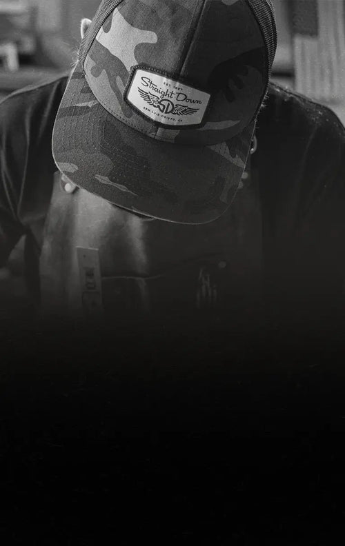 Black and white photo of a craftsman working in the woodshop wearing a camo hat. With an american flag hanging on the wall above his shoulder. 