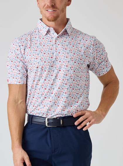 Grillin' Polo in White, on model front view featuring a white base with an all-over scattered  print of American flags, watermelon, hot dogs, grills, and fireworks in red and blue tones, styled with navy pants and a navy belt