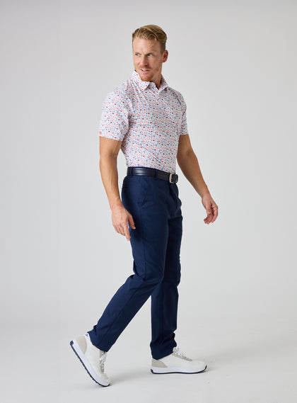 Grillin' Polo in White, full body on model side view featuring a white base with an all-over scattered print in red and blue tones, styled with navy pants, a navy belt, and white golf shoes