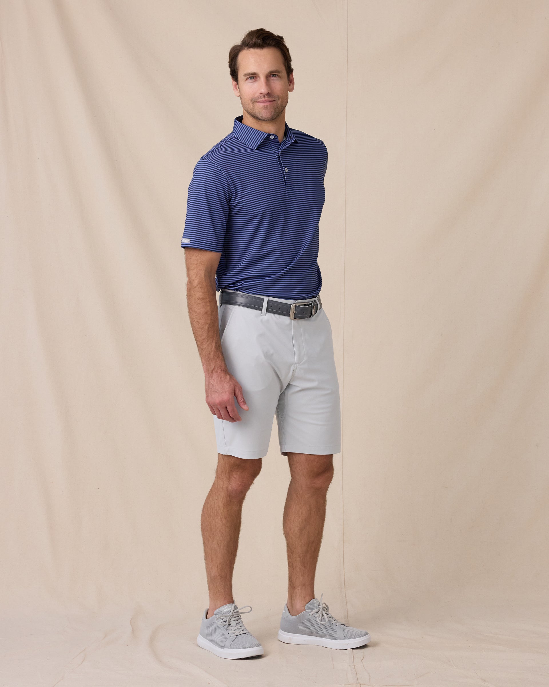 Men’s striped tidal blue polo styled with silver shorts, grey belt, and grey sneakers