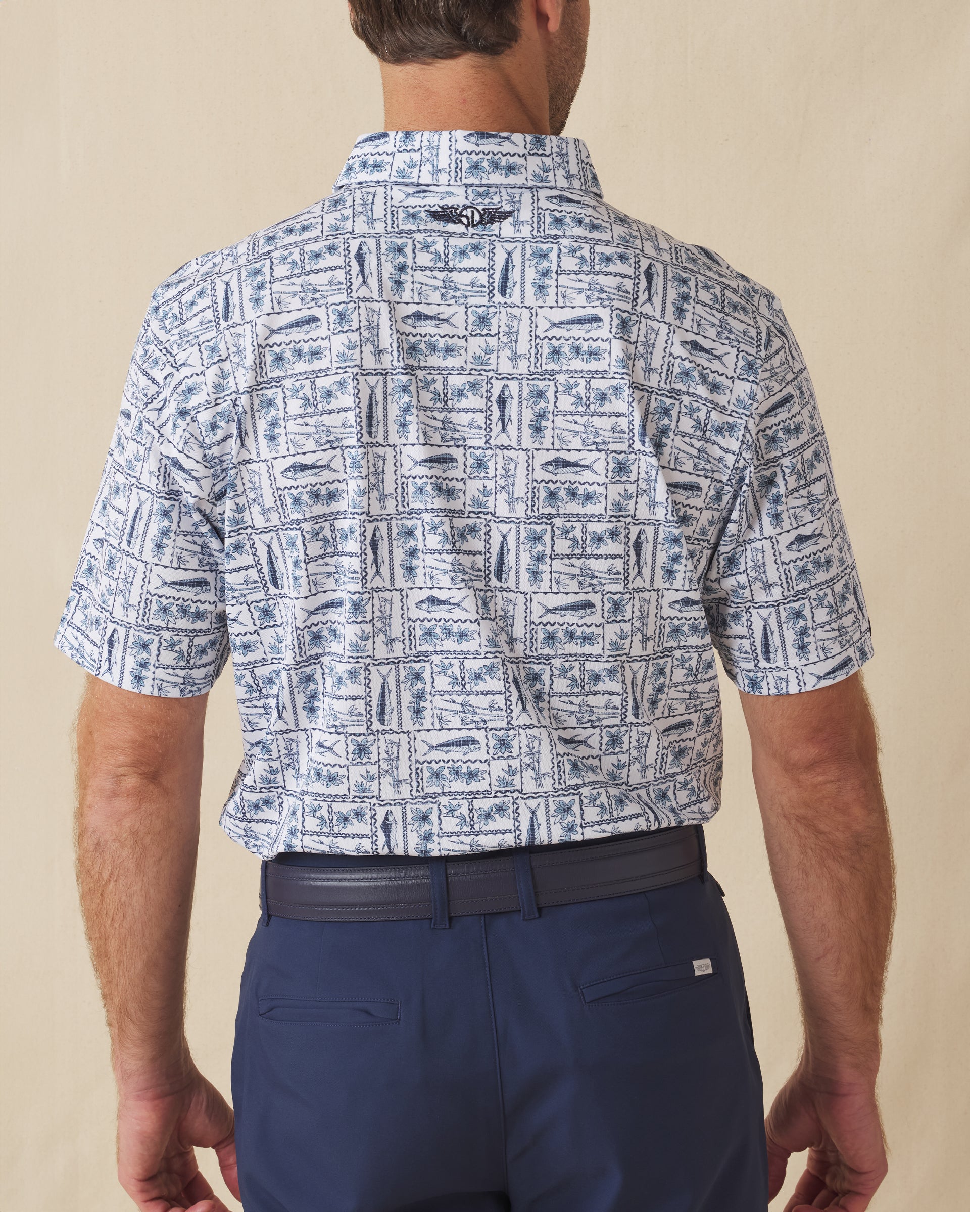 Men's Dorado Polo in Coastal, back view on model featuring a white base with a navy and blue tiled fish, floral, and bamboo stamp print, with Straight Down logo embroidered at upper back
