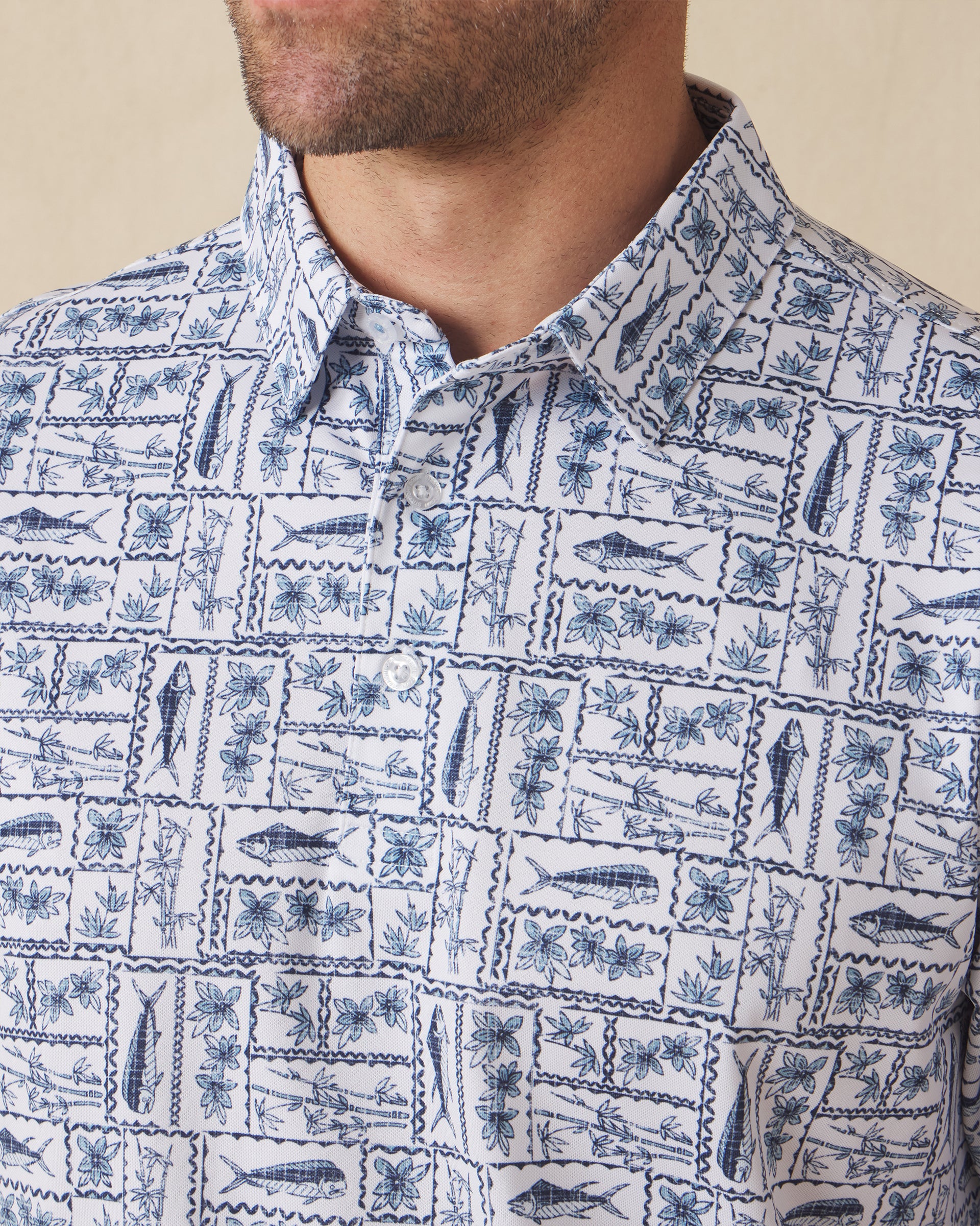 Men's Dorado Polo in Coastal, close-up front view on model highlighting the navy and blue tiled fish, floral, and bamboo stamp print on a white base