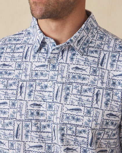 Men's Dorado Polo in Coastal, close-up front view on model highlighting the navy and blue tiled fish, floral, and bamboo stamp print on a white base