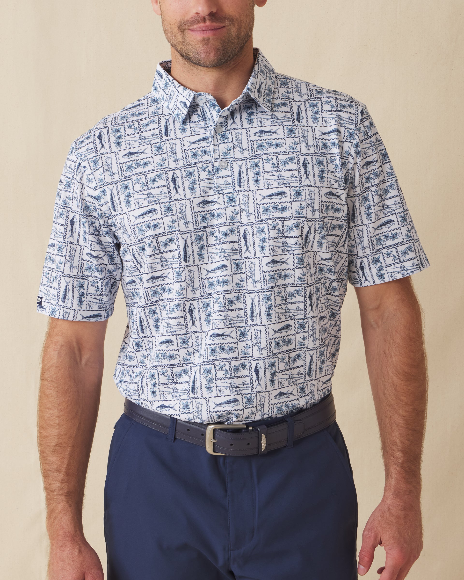 Men's Dorado Polo in Coastal, front view on model featuring a white base with a navy and blue tiled fish, floral, and bamboo stamp print, paired with navy pants and a dark leather belt