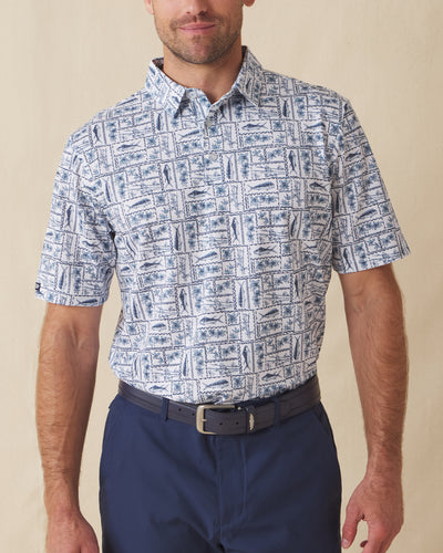Men's Dorado Polo in Coastal, front view on model featuring a white base with a navy and blue tiled fish, floral, and bamboo stamp print, paired with navy pants and a dark leather belt