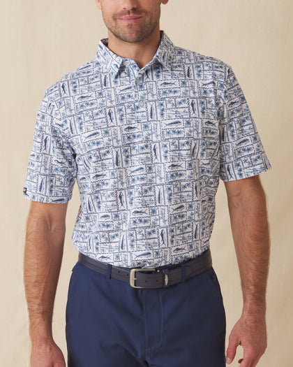 Men's Dorado Polo in Coastal, front view on model featuring a white base with a navy and blue tiled fish, floral, and bamboo stamp print, paired with navy pants and a dark leather belt