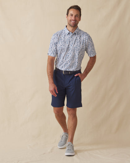 Men's Dorado Polo in Coastal, full length front view on model featuring a white base with a navy and blue tiled fish, floral, and bamboo stamp print, paired with navy shorts, a dark leather belt, and gray shoes