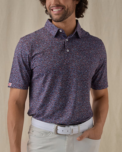 Men's American Muscle Polo in New Indigo, front view on model featuring a navy, burgundy, and sky blue classic car novelty print, paired with light gray pants and a white belt, with an American flag woven label at the sleeve