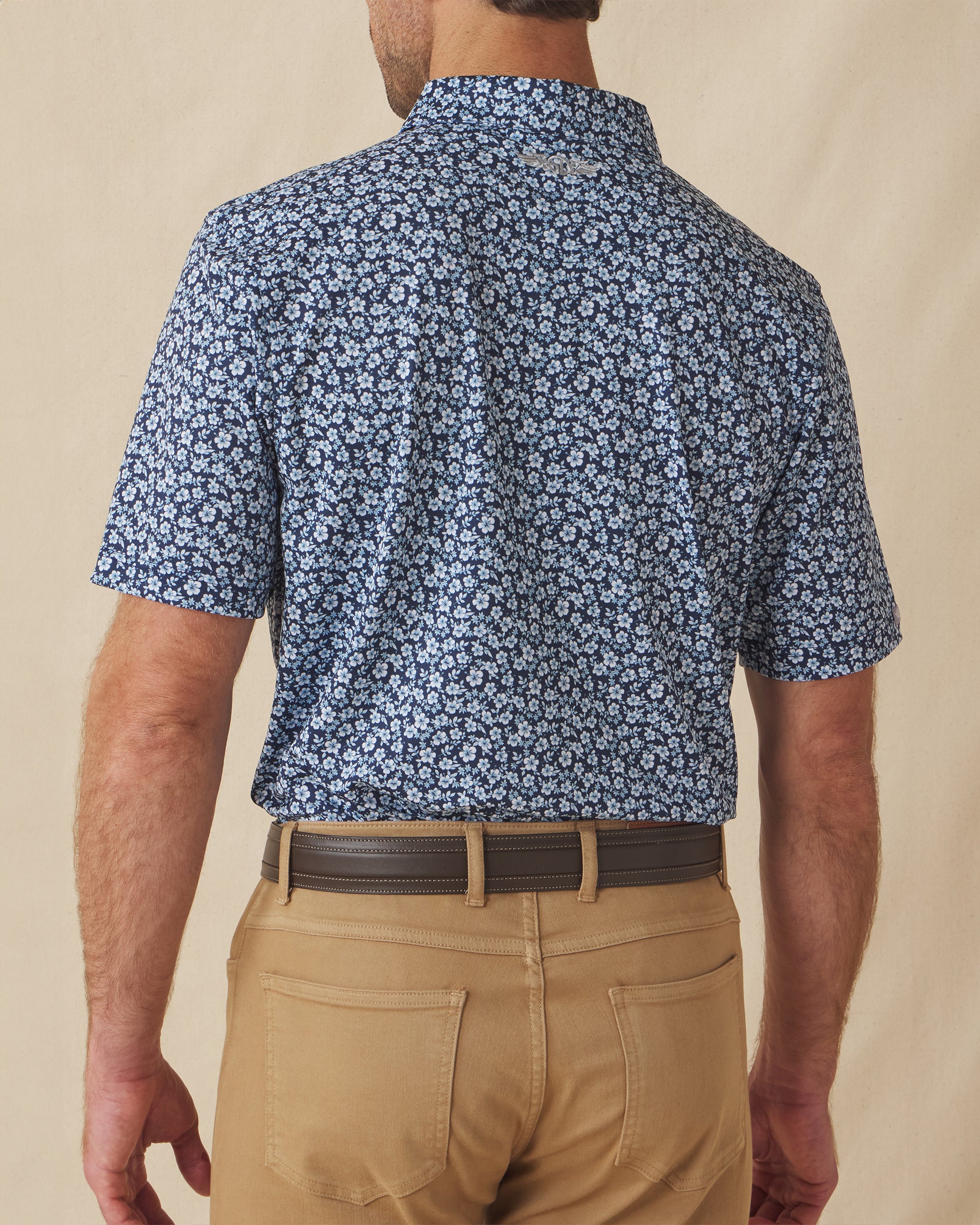 Men's Don Ho Polo in New Indigo, back view on model featuring a navy base with an all-over light blue floral print, with Straight Down logo embroidered at upper back, paired with khaki pants