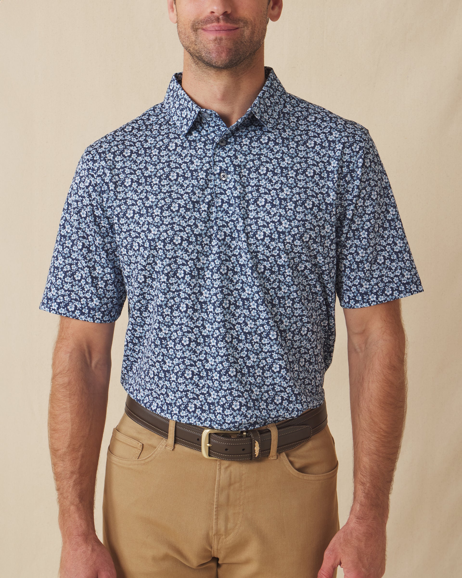 Men's Don Ho Polo in New Indigo, front view on model featuring a navy base with an all-over light blue floral print, paired with khaki pants and a dark leather belt