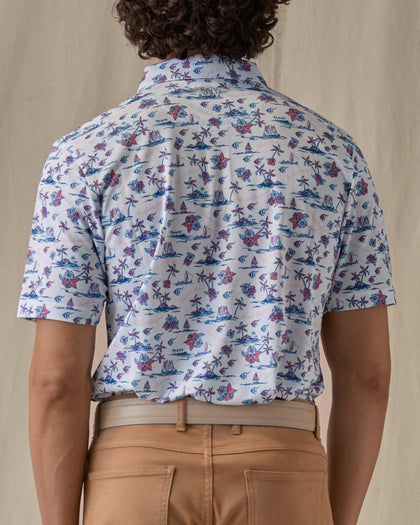 Men's Island Paradise Polo in Cool Blue, back view on model featuring an all-over tropical print with palm trees, hibiscus flowers, sailboats, and tropical fish and blue on a white body, worn with tan pants