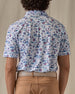 Men's Island Paradise Polo in Cool Blue, back view on model featuring an all-over tropical print with palm trees, hibiscus flowers, sailboats, and tropical fish and blue on a white body, worn with tan pants