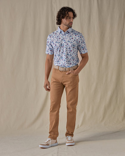 Men's Island Paradise Polo in Cool Blue, full front view on model featuring an all-over tropical print with palm trees, hibiscus flowers, sailboats, and tropical fish and blue on a white body, worn with tan pants, a tan belt, and tan and white golf shoes